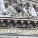 Detail of the carved stone facade of the New York Stock Exchange building in New York, with its gold-lettered STOCK EXCHANGE inscription below classical sculptures.