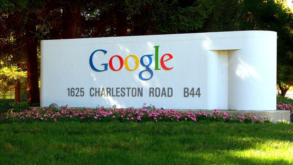 The colourful Google logo on the white entrance sign at 1625 Charleston Road, Google's Mountain View campus, with pink flowers and grass in front.