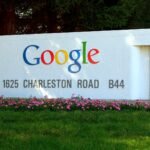 The colourful Google logo on the white entrance sign at 1625 Charleston Road, Google's Mountain View campus, with pink flowers and grass in front.