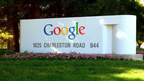 The colourful Google logo on the white entrance sign at 1625 Charleston Road, Google's Mountain View campus, with pink flowers and grass in front.