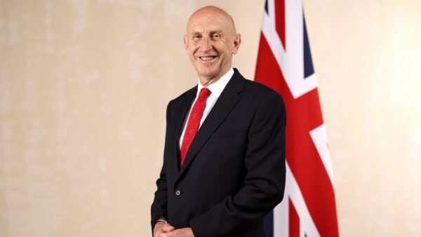 Chancellor John Healey in a dark suit and red tie standing beside a Union flag in his official cabinet portrait.