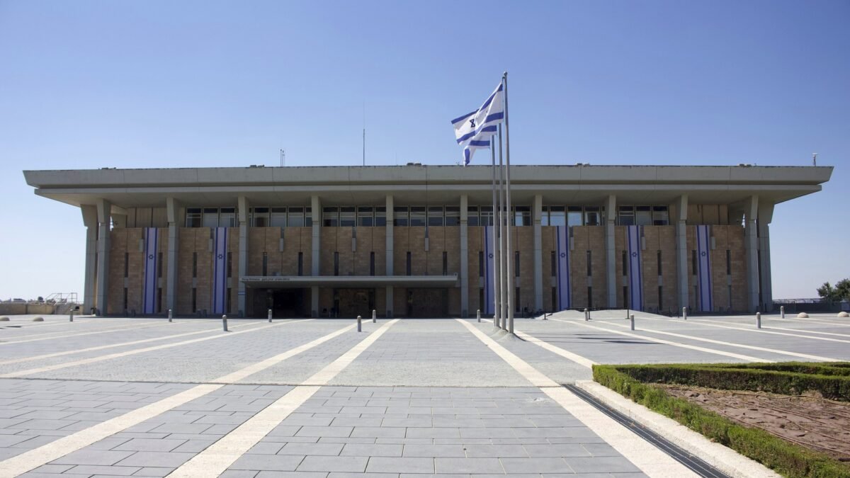 The Knesset building in Jerusalem, with Israeli flags flying in front of the parliament's stone facade.