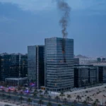 Smoke rises from an upper floor of a modern glass office complex at dusk in a Gulf oil city, with emergency vehicles below.