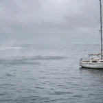 A small white sailing yacht on the grey Baltic Sea with gas bubbles breaking on the water surface under an overcast sky