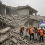 Rescue workers in orange vests clearing rubble at the collapsed Al-Khair Education Centre school in Peshawar, Pakistan, with an ambulance nearby.