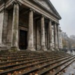 Stone facade and steps of a London courthouse on a grey autumn morning, with journalists and camera tripods gathered outside during Russell Brand's trial.