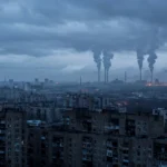 Kyiv city skyline at dawn under heavy cloud, with tall smoke columns rising from power plant chimneys on the outskirts and dark apartment blocks in the foreground