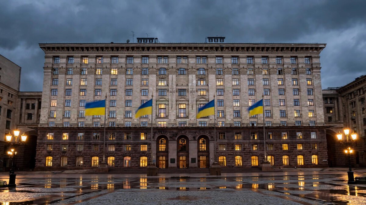 A grand government ministry building in Kyiv at dusk, with large Ukrainian blue-and-yellow flags flying from tall flagpoles in front