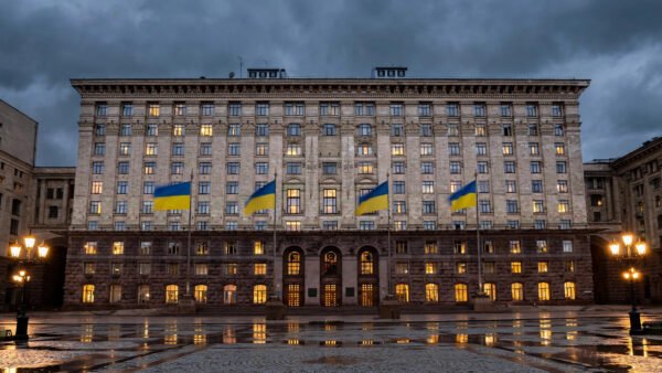 A grand government ministry building in Kyiv at dusk, with large Ukrainian blue-and-yellow flags flying from tall flagpoles in front