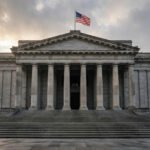 Neoclassical central bank building facade with tall stone columns and an American flag under a dramatic morning sky, illustrating the Federal Reserve