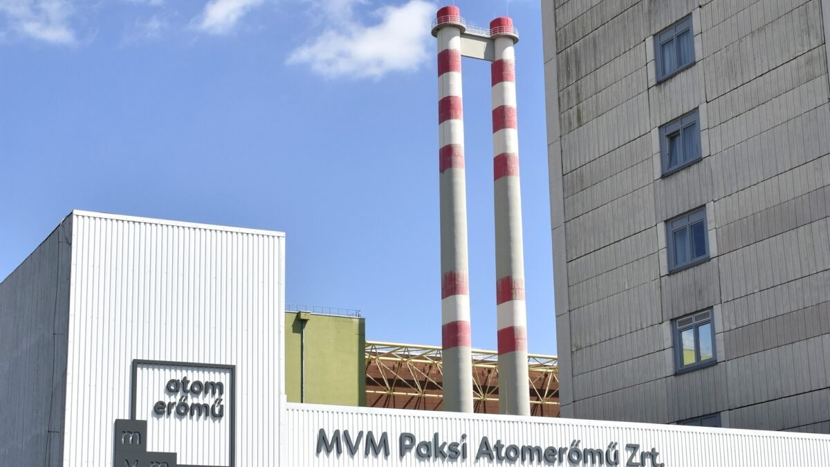 The main entrance of the Paks Nuclear Power Plant in Hungary, with MVM Paksi Atomeromu Zrt. signage, an atom eromu sign, and twin red-and-white chimneys against a blue sky.