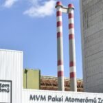 The main entrance of the Paks Nuclear Power Plant in Hungary, with MVM Paksi Atomeromu Zrt. signage, an atom eromu sign, and twin red-and-white chimneys against a blue sky.