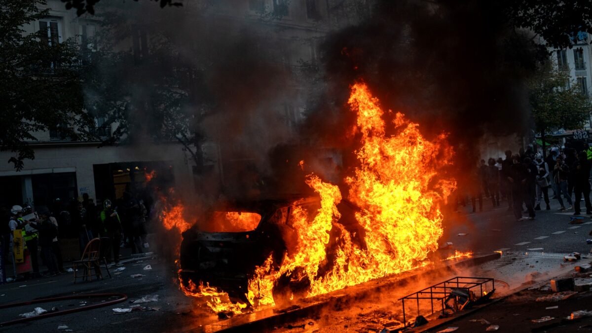 A car burns on a Paris street during the 17 October 2026 riots as demonstrators watch from a distance
