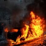 A car burns on a Paris street during the 17 October 2026 riots as demonstrators watch from a distance