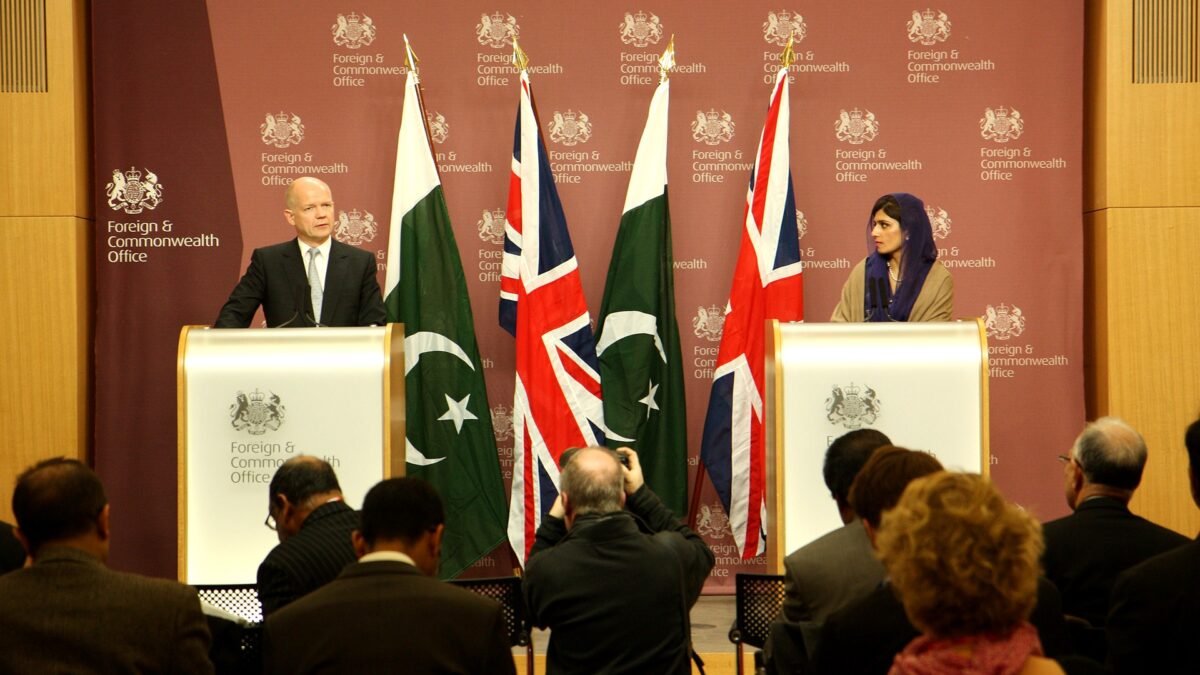A press photographer documents a press conference featuring Pakistan's foreign minister, with Pakistani flags behind the podium.