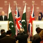 A press photographer documents a press conference featuring Pakistan's foreign minister, with Pakistani flags behind the podium.
