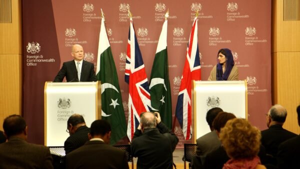 A press photographer documents a press conference featuring Pakistan's foreign minister, with Pakistani flags behind the podium.