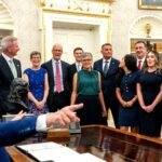 US President Donald Trump speaking in the Oval Office, with guests standing behind him.