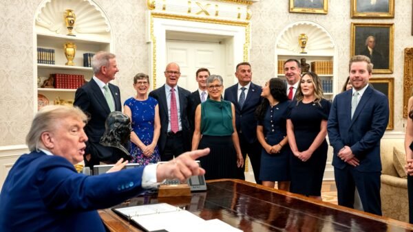 US President Donald Trump speaking in the Oval Office, with guests standing behind him.