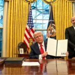 President Donald Trump holds up a signed executive order at the Resolute Desk in the Oval Office, flanked by aides and the US and presidential flags.