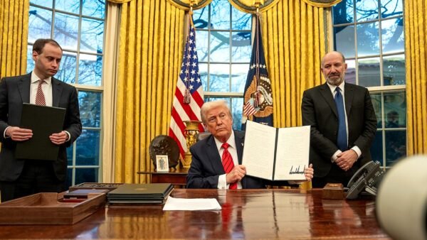President Donald Trump holds up a signed executive order at the Resolute Desk in the Oval Office, flanked by aides and the US and presidential flags.