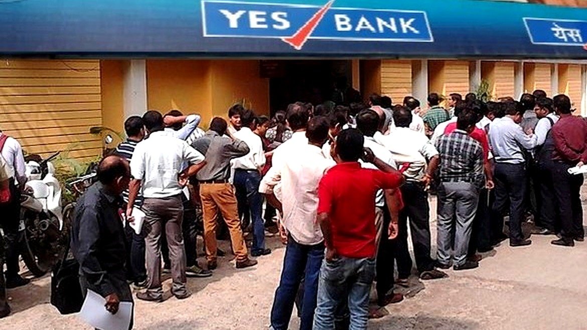 Depositors queue outside a Yes Bank branch, with the bank's blue-and-red YES BANK sign visible above the entrance.